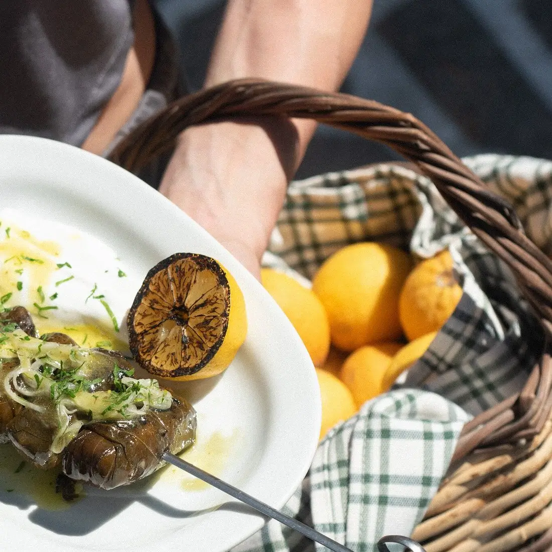 Grilled lemon and skewered cooked eggplant on a white plate held over a basket of lemons.