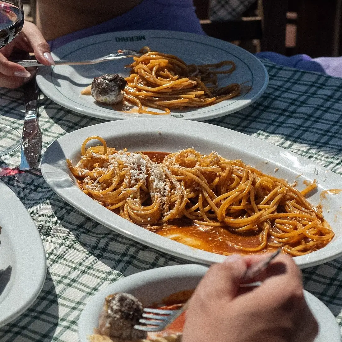 Plates of spaghetti with tomato sauce and meatballs on a checkered tablecloth with people eating.