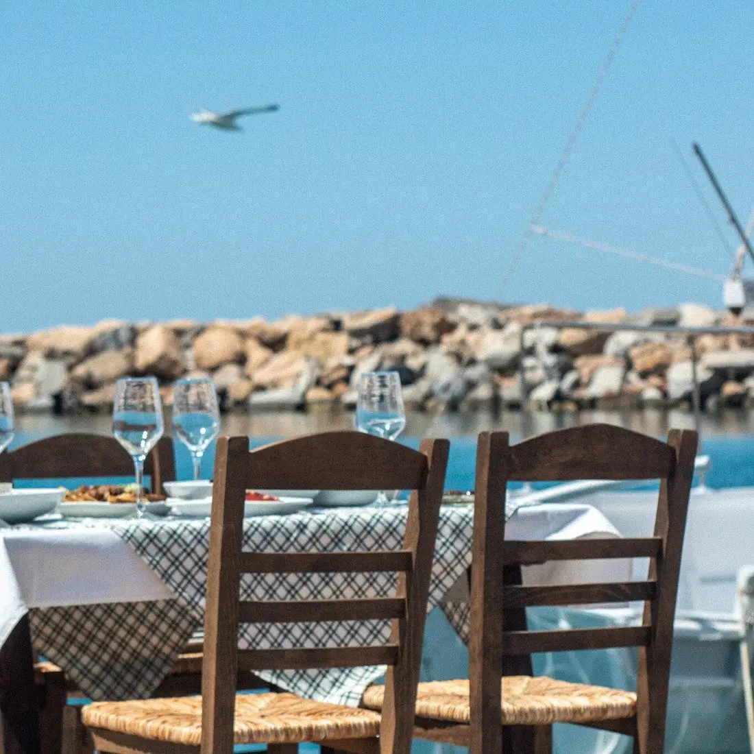 Outdoor dining table with checkered cloth and empty wine glasses by water and rocky shore.