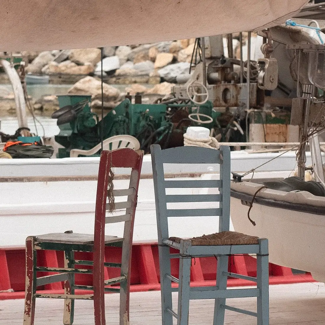 Two weathered wooden chairs, one red and one blue, placed on a boat deck near harbor equipment.