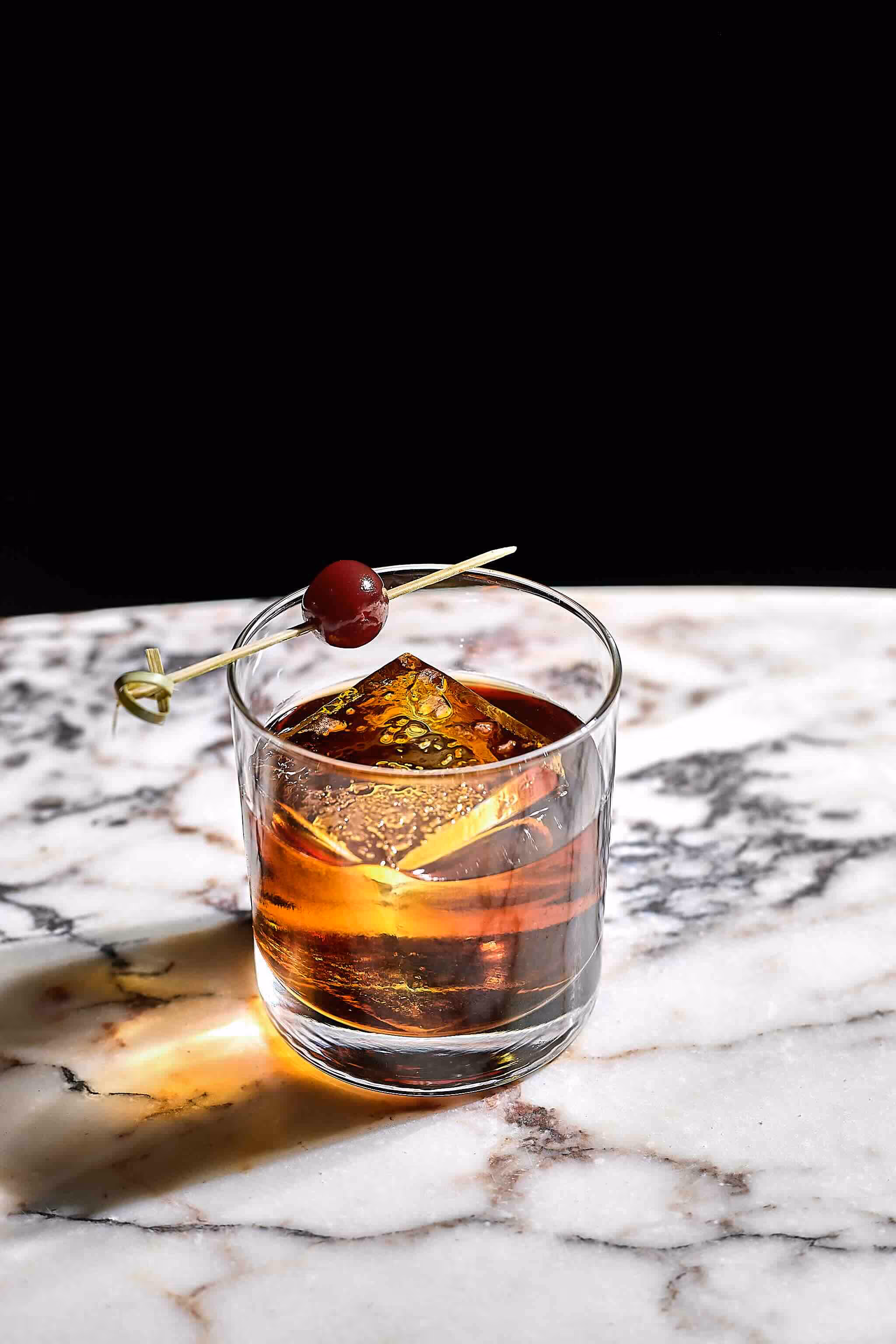 Whiskey cocktail with large ice cube and cherry garnish on a marble surface with black background.