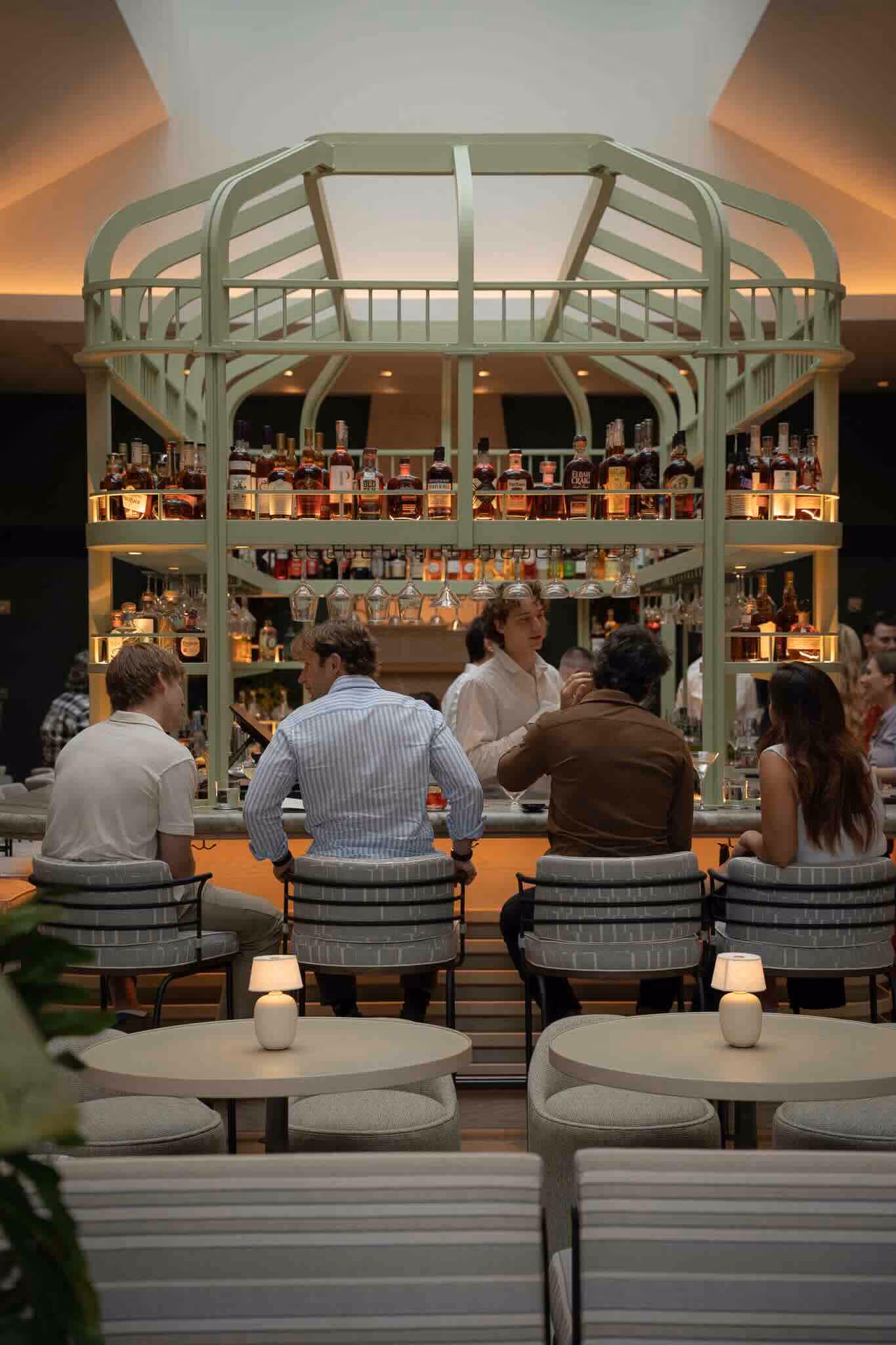 Four people seated at a bar counter with bottles and glasses on shelves above them.