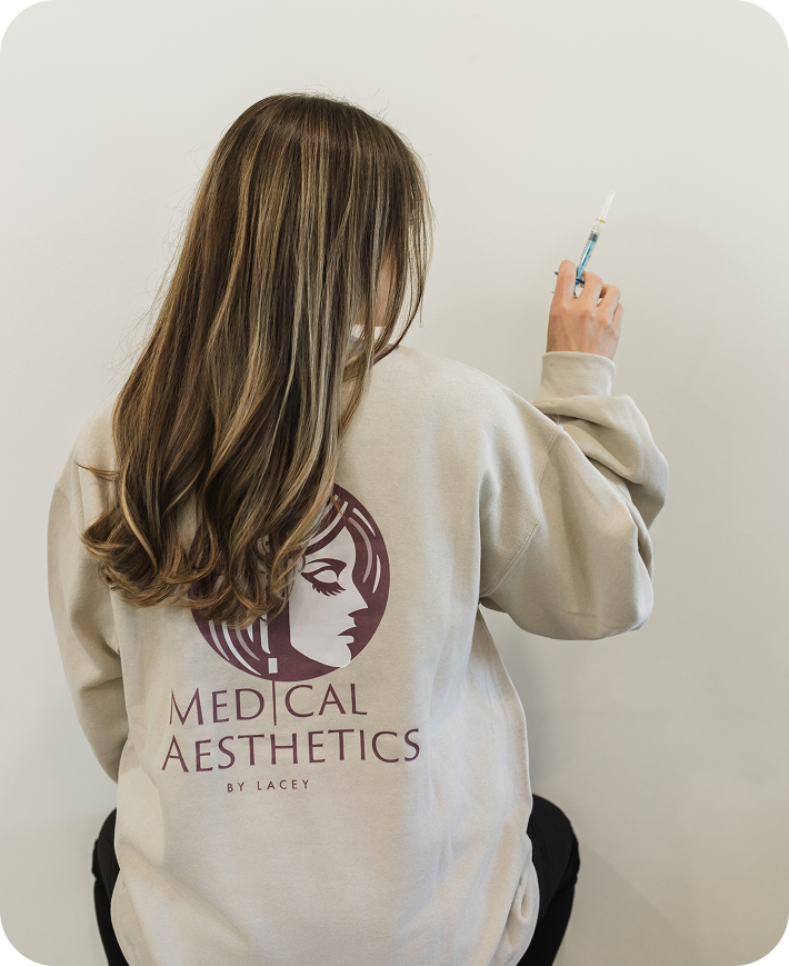 Person with long hair in Medical Aesthetics by Lacey sweatshirt holding a syringe against a white wall.