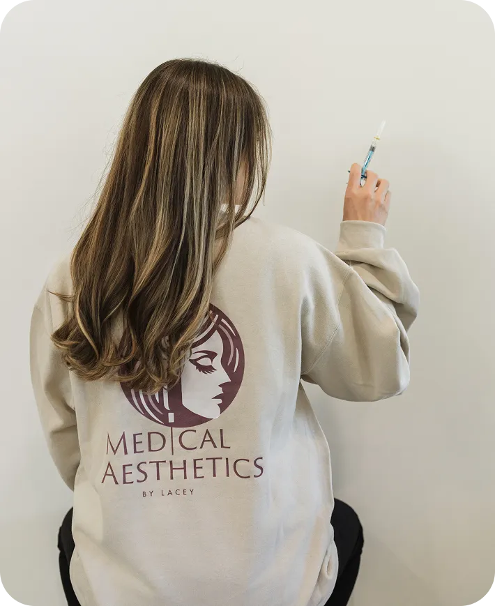 Person with long hair in Medical Aesthetics by Lacey sweatshirt holding a syringe against a white wall.