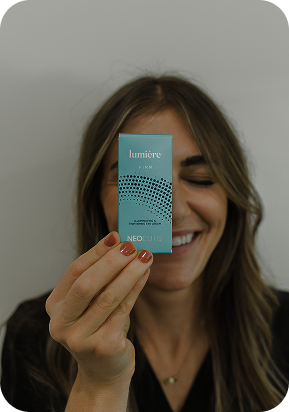 Smiling woman holding blue Lumière PRM Neocutis box near her face against gray wall.