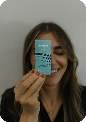 Smiling woman holding blue Lumière PRM Neocutis box near her face against gray wall.