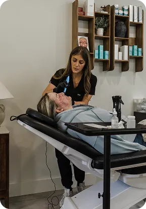Woman receiving a facial treatment from a technician in a spa room with skincare products on shelves.
