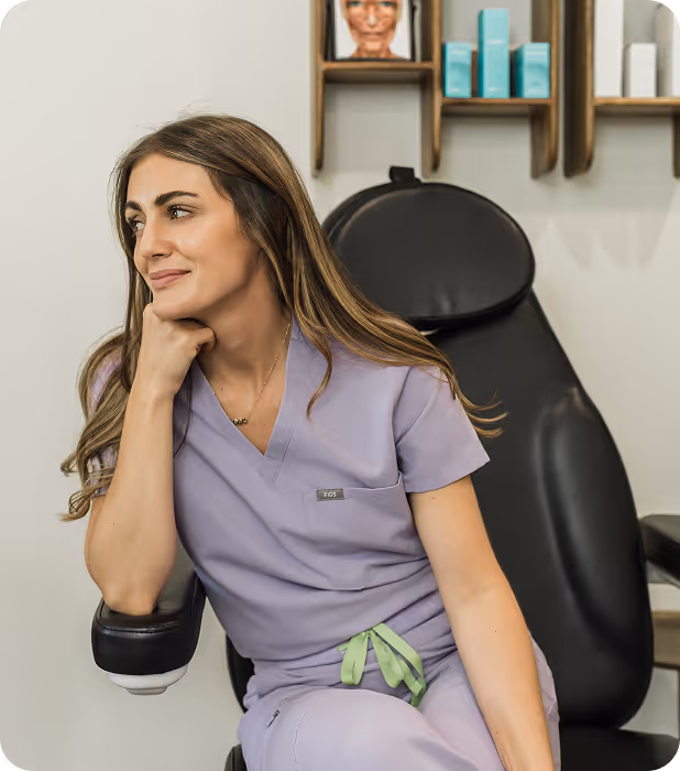 Woman in lavender scrub uniform sitting on a black chair in a medical or skincare office.