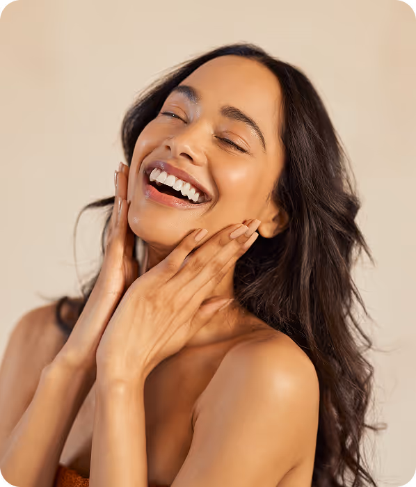 Smiling woman with long dark hair, eyes closed, gently touching her face with both hands.