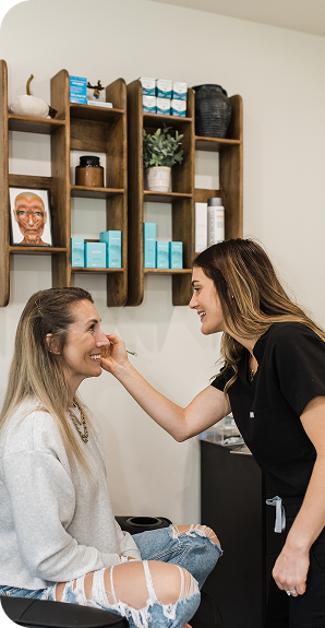 Woman applying makeup to another smiling woman sitting in a chair inside a beauty salon.