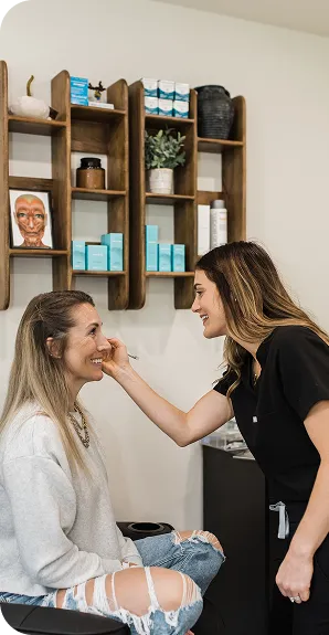 Woman applying makeup to another smiling woman sitting in a chair inside a beauty salon.