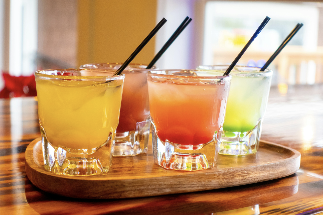 Four colorful cocktails with black straws on a wooden tray on a polished wooden table.