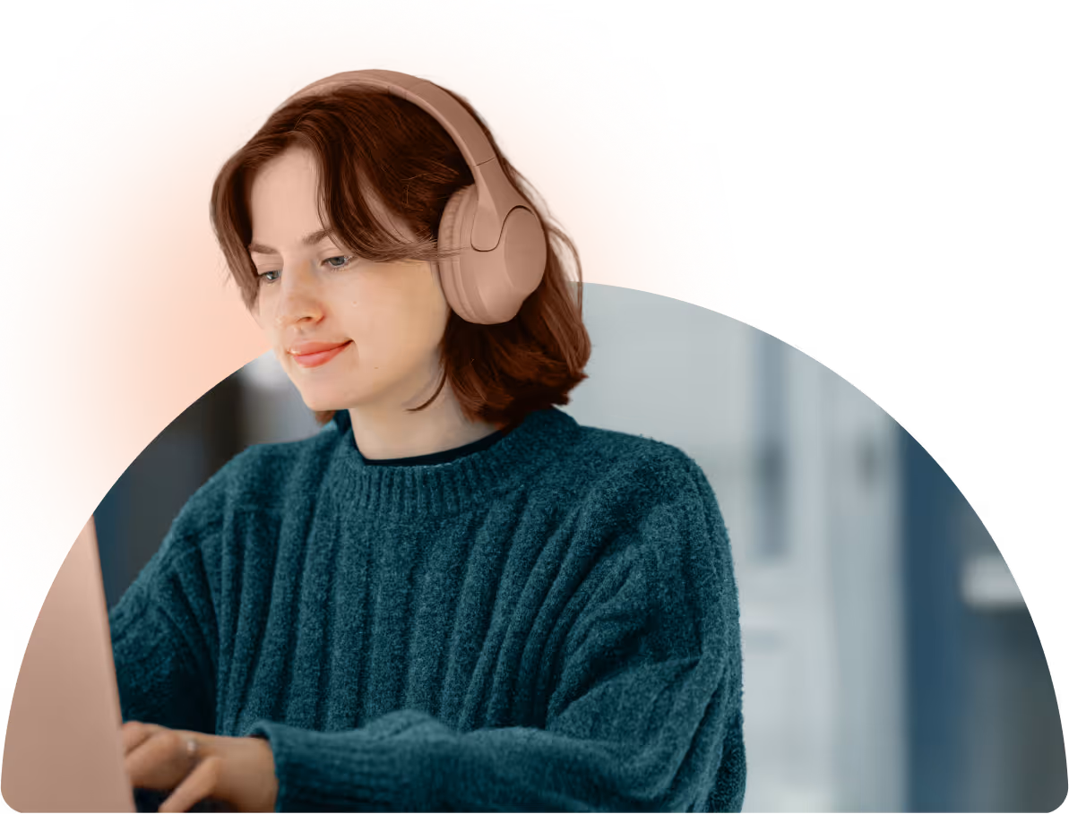 Young woman with brown hair wearing pink headphones and a teal sweater working on a laptop.