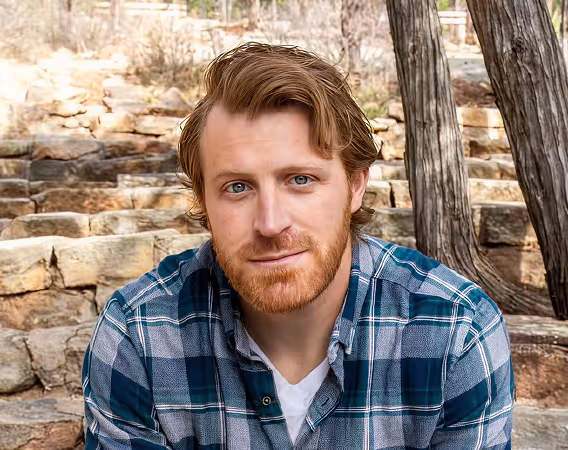 Man with red hair and beard wearing blue plaid shirt sitting outdoors on stone steps.