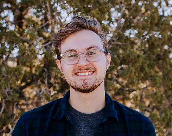 Young man with glasses and beard smiling outdoors with blurred tree background.
