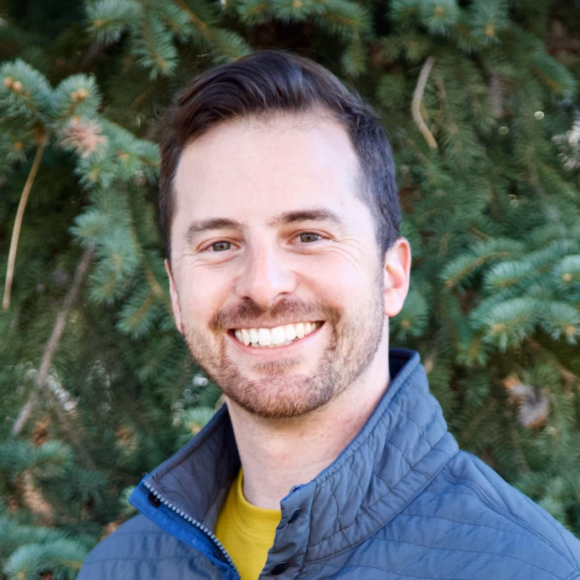 Smiling man with short dark hair and beard wearing a blue jacket and yellow shirt, standing outdoors with green pine tree background.