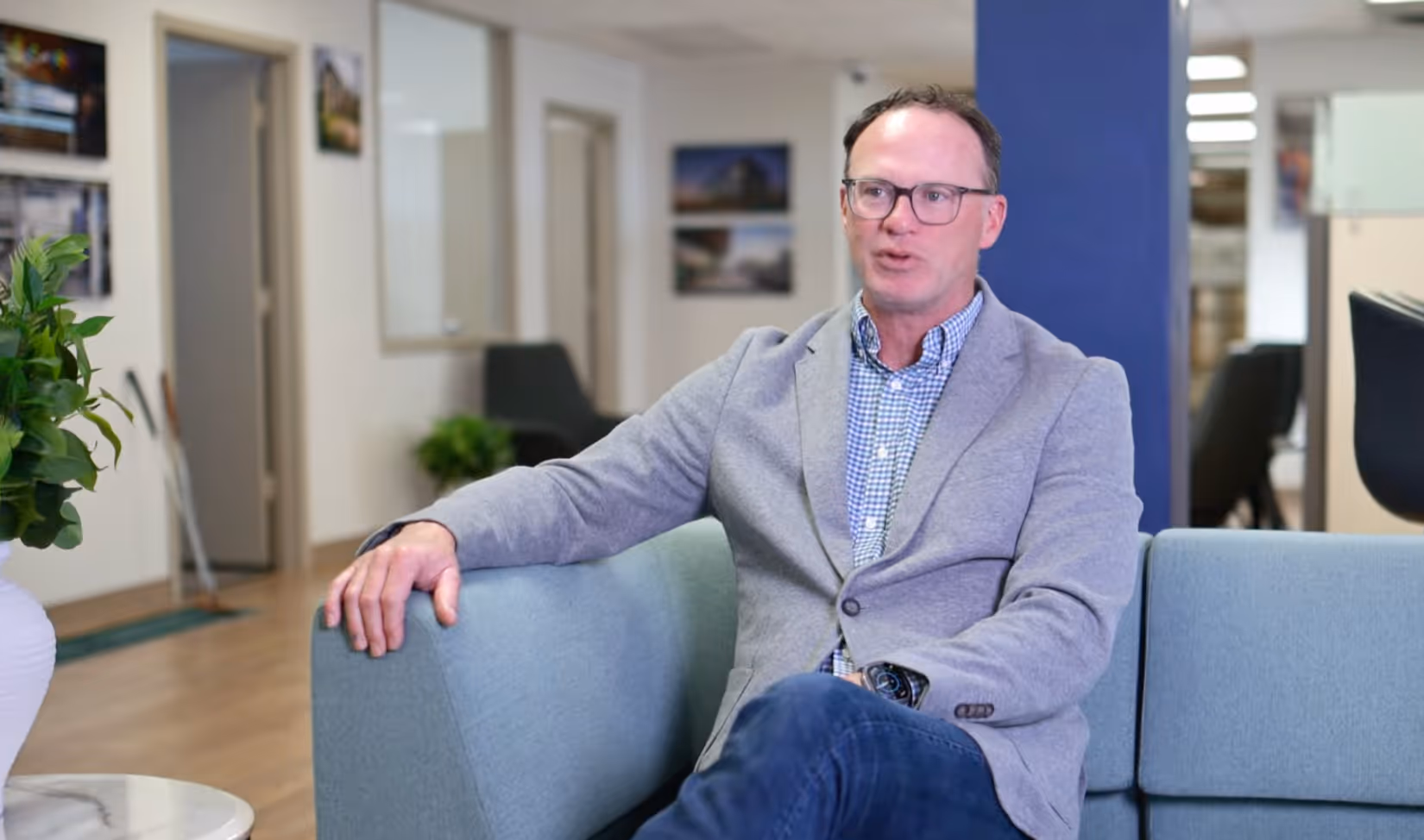Man wearing glasses and gray blazer sitting on a light blue sofa in a modern office setting.