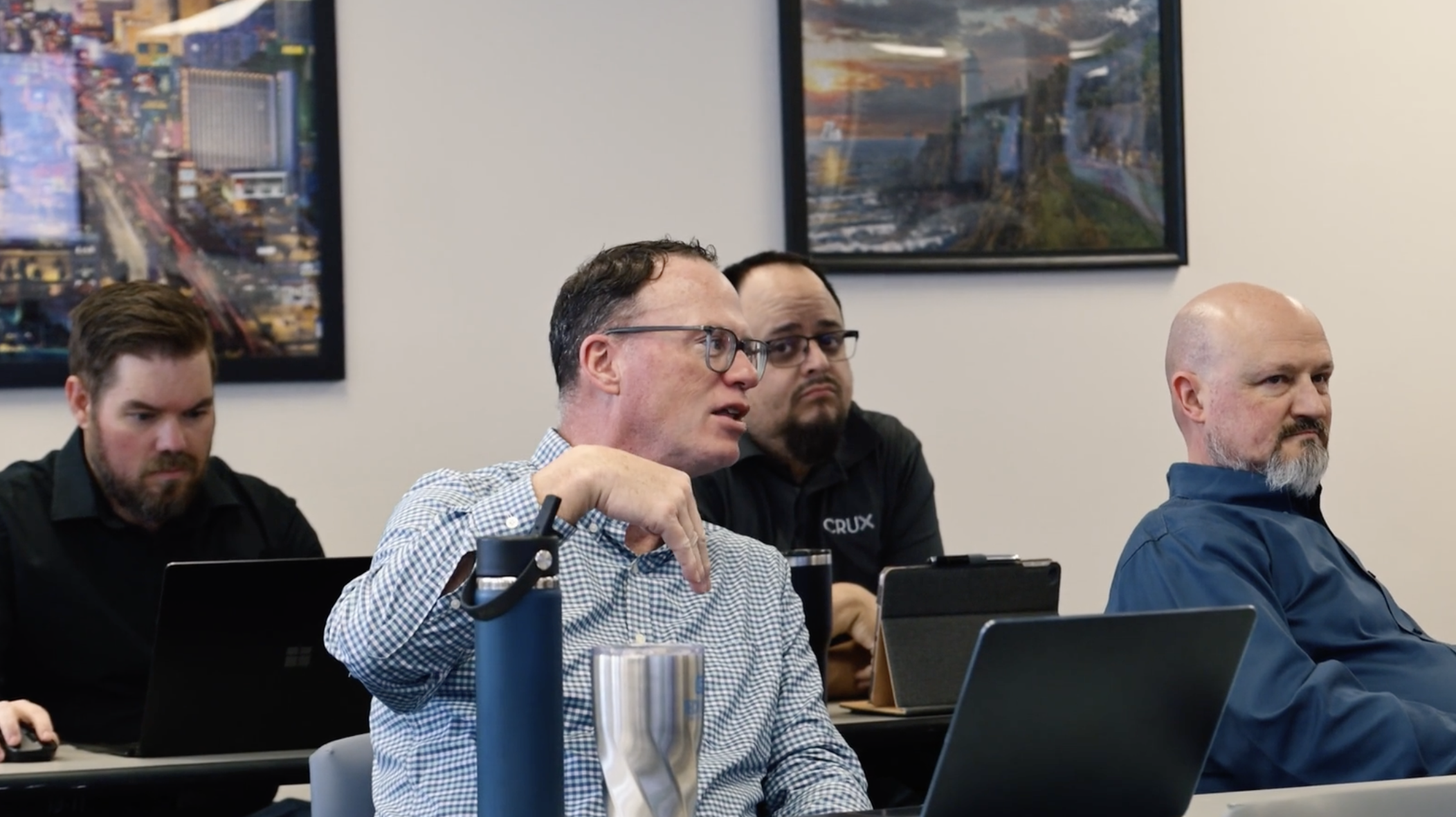 Four men in a meeting room with laptops, one speaking and others listening attentively.