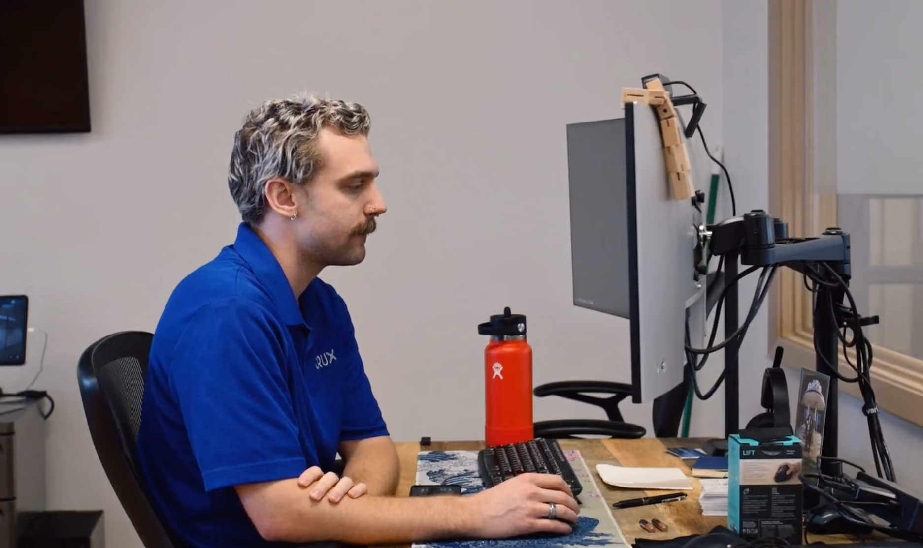 Man with mustache and curly hair working on a computer at a desk with a red water bottle.