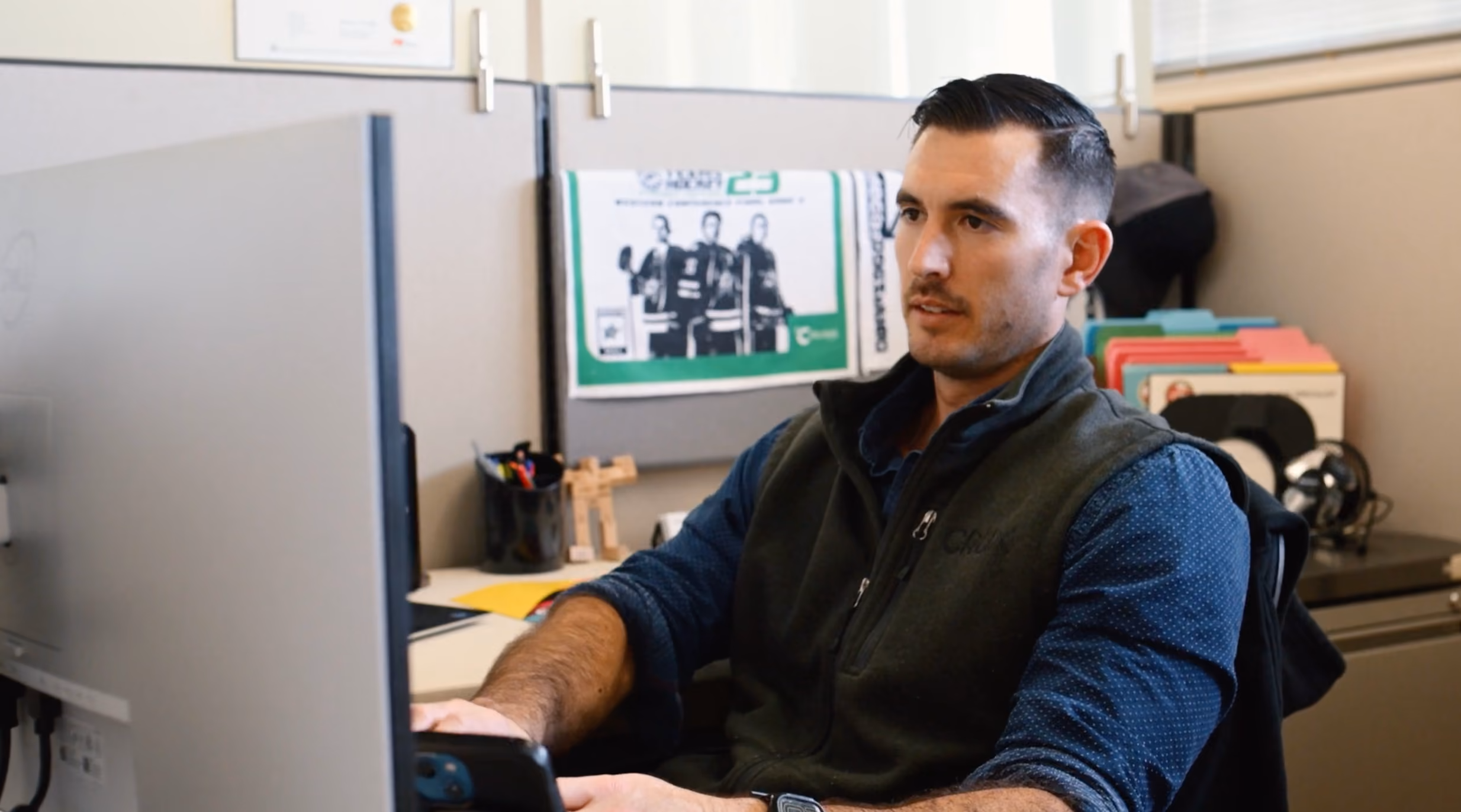 Man with dark hair and mustache working at computer desk in office cubicle with files behind him.