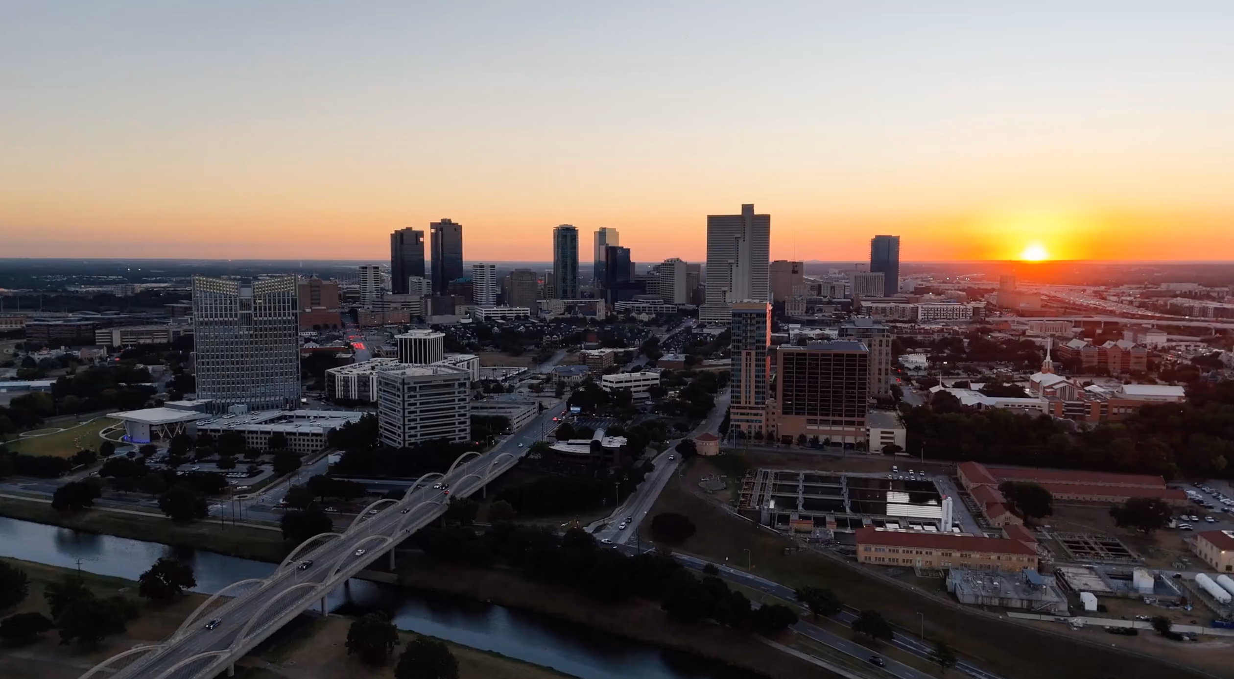 Sunset over a city skyline with a river, bridge, and modern high-rise buildings.