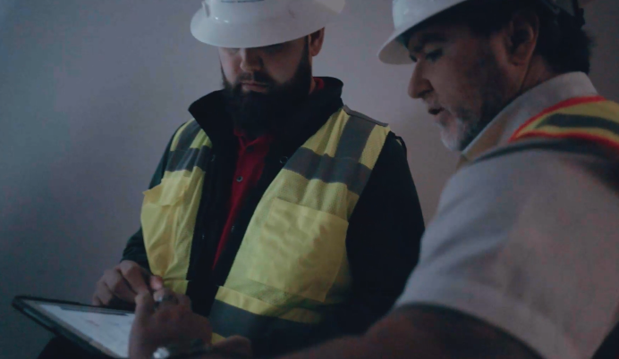 Two construction workers in helmets and safety vests reviewing plans on a clipboard.