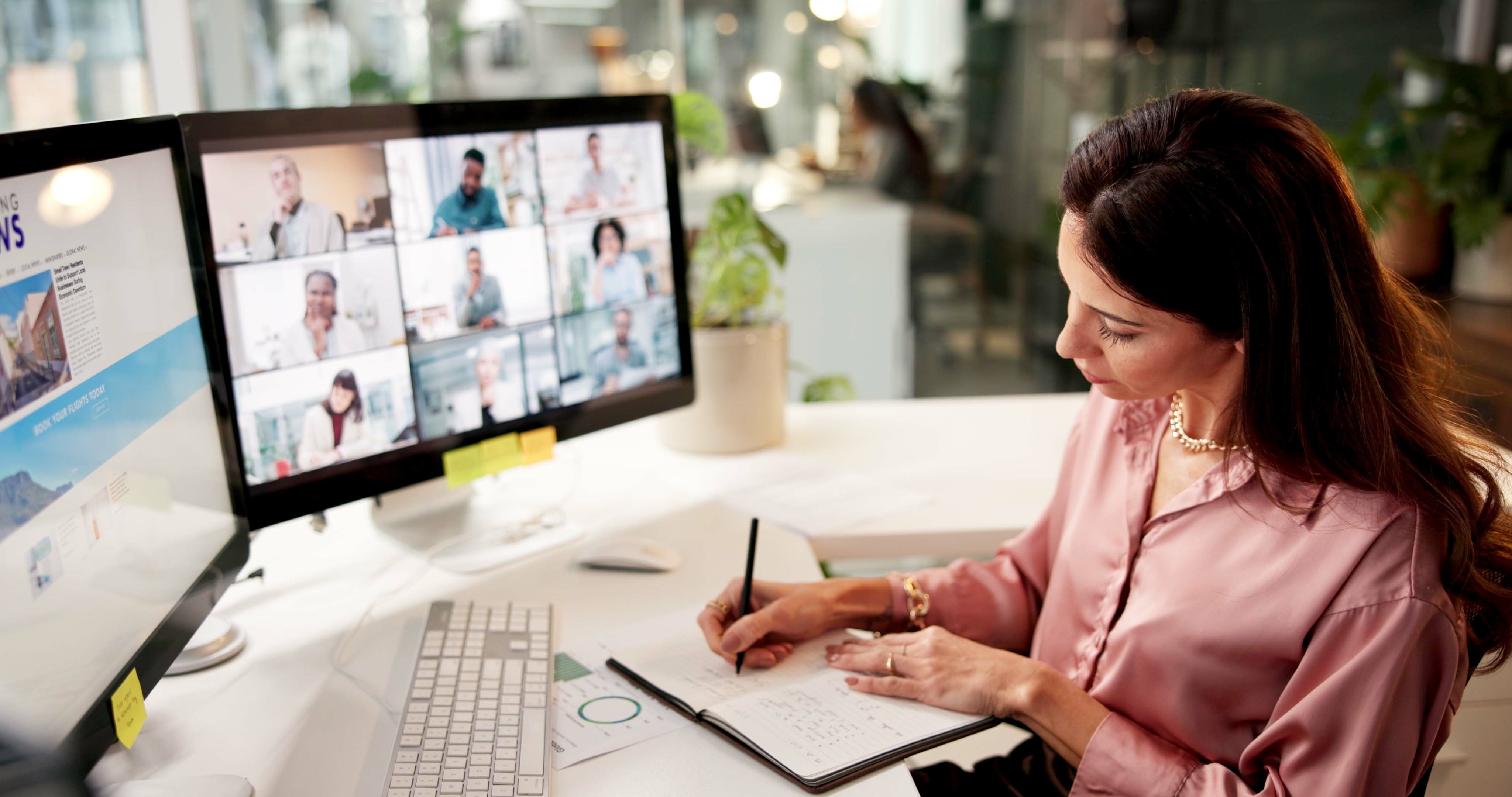 Woman in pink blouse writing notes while attending a virtual meeting on dual computer monitors.