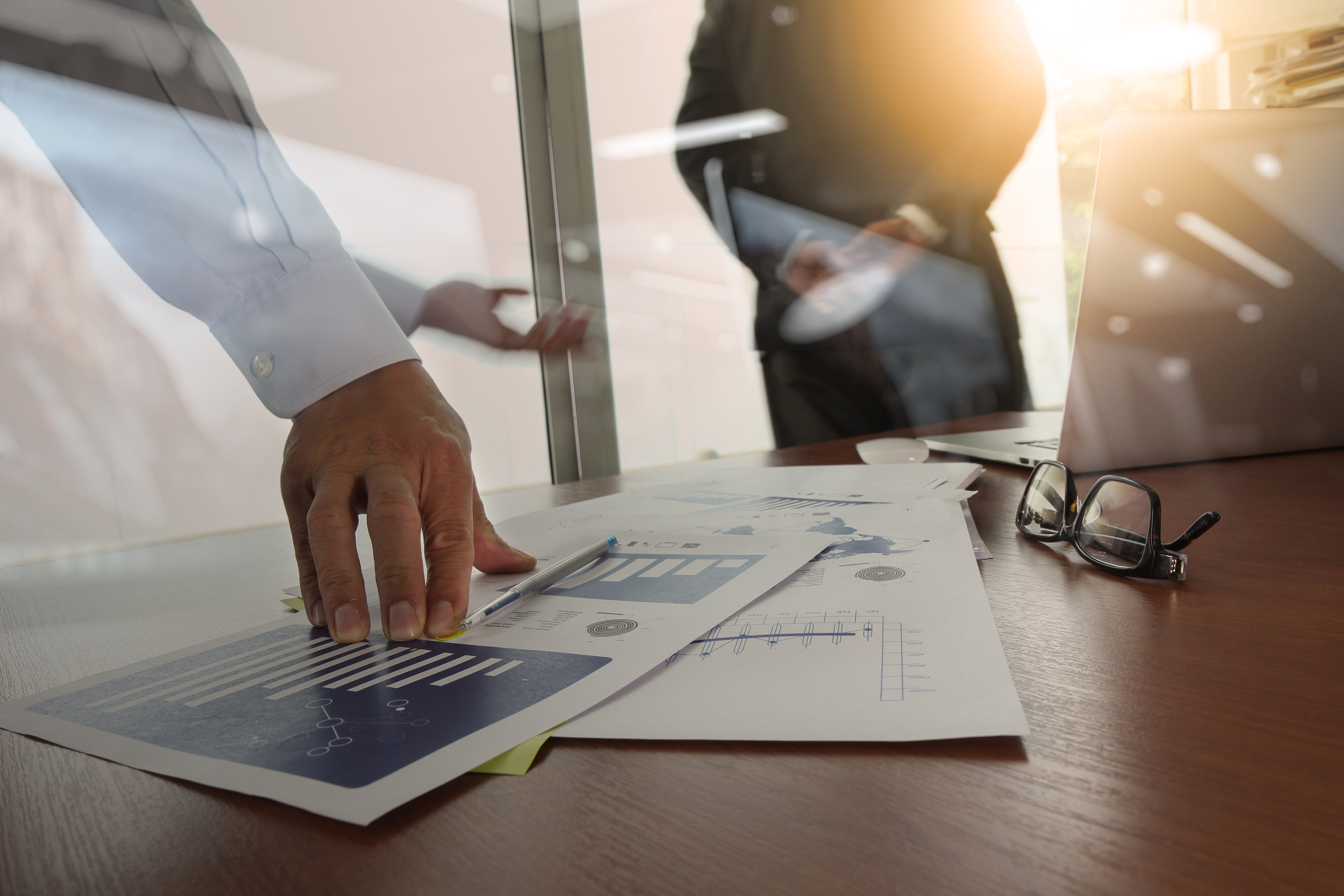 Hand pointing at business charts on desk with glasses and laptop nearby in a meeting.