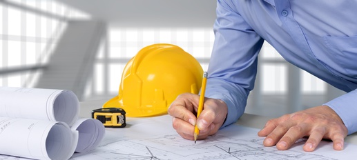 Person in blue shirt drawing on architectural plans with yellow hard hat and tape measure nearby.