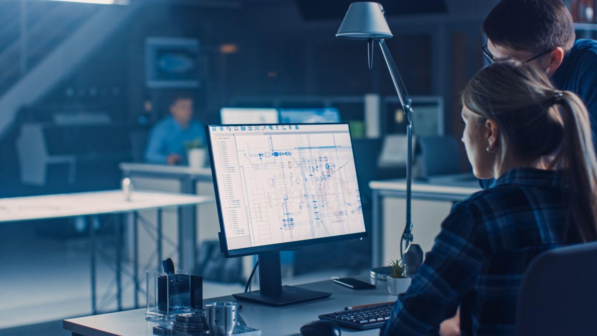 Two people reviewing technical blueprints displayed on a computer monitor in a modern office.