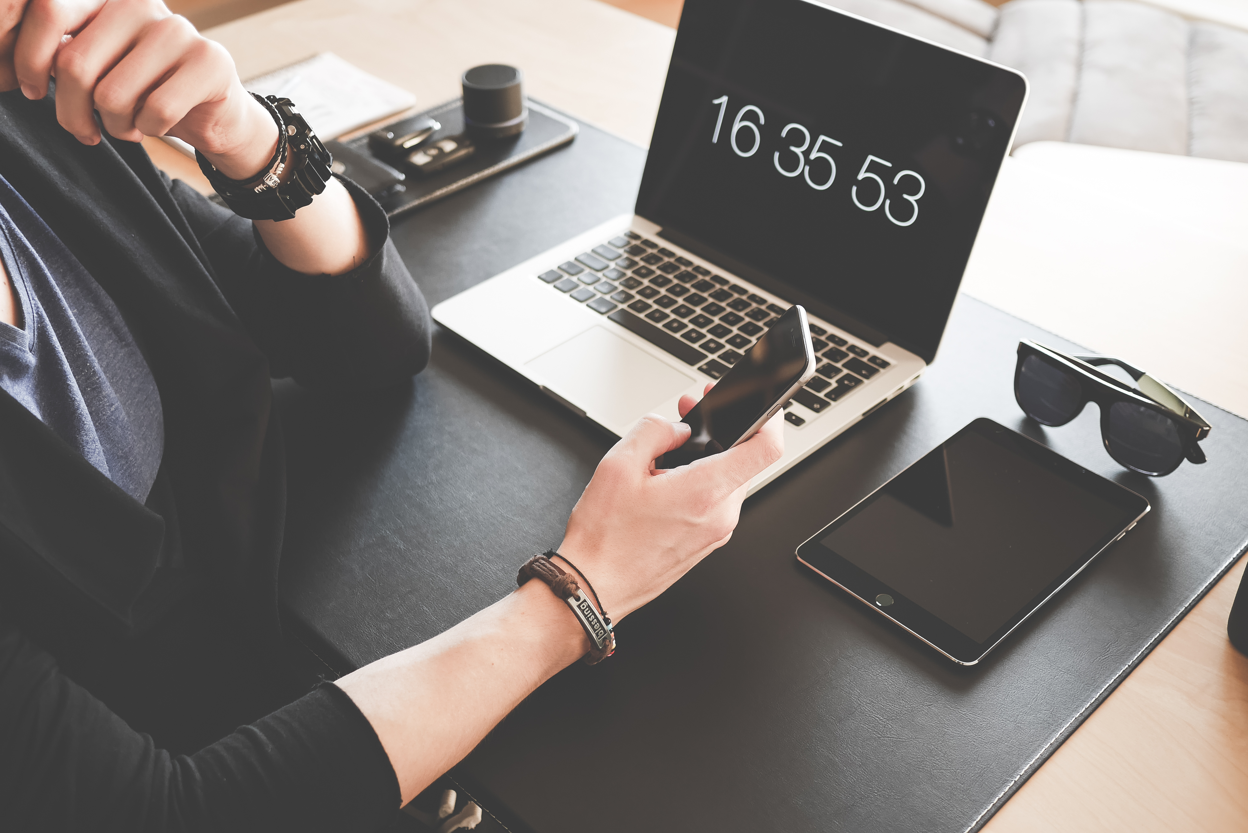 Person holding smartphone near a laptop showing digital clock, tablet, and sunglasses on desk.