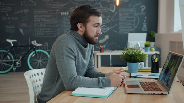 Young bearded man is making internet call talking to friends online from open space office looking at laptop screen. People and technology concept.
