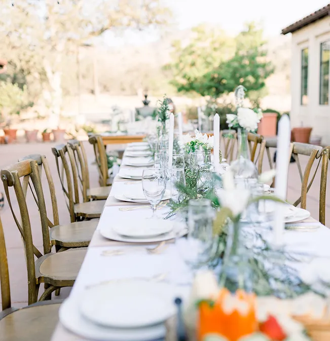 Long outdoor banquet table with taper candles, white florals, and cross-back chairs beside a stucco building
