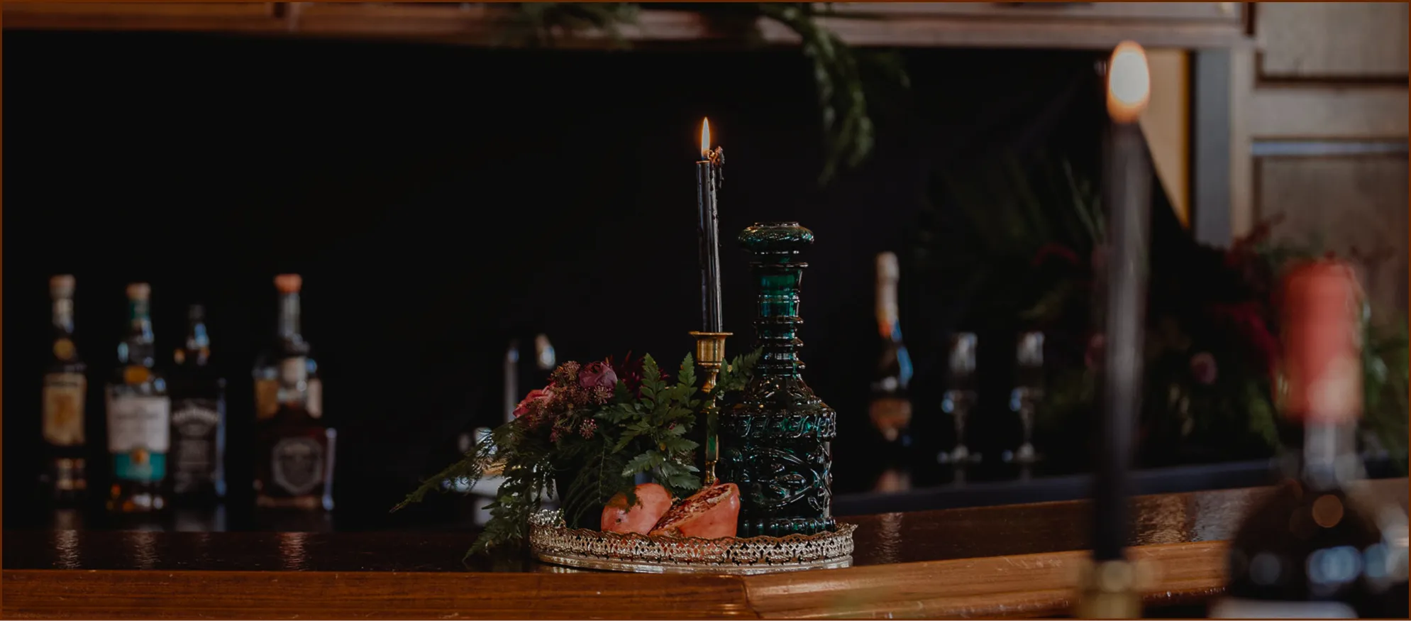 Candlelit bar vignette with a green glass decanter, flowers, and bottles in the background