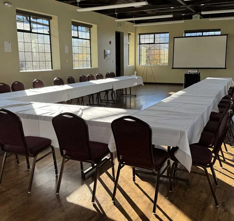 Sunlit meeting room with U-shaped white-clothed tables and chairs facing a projection screen