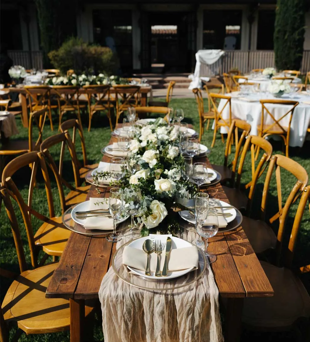Rustic outdoor wedding reception with wooden farm tables, crossback chairs, and white rose centerpieces