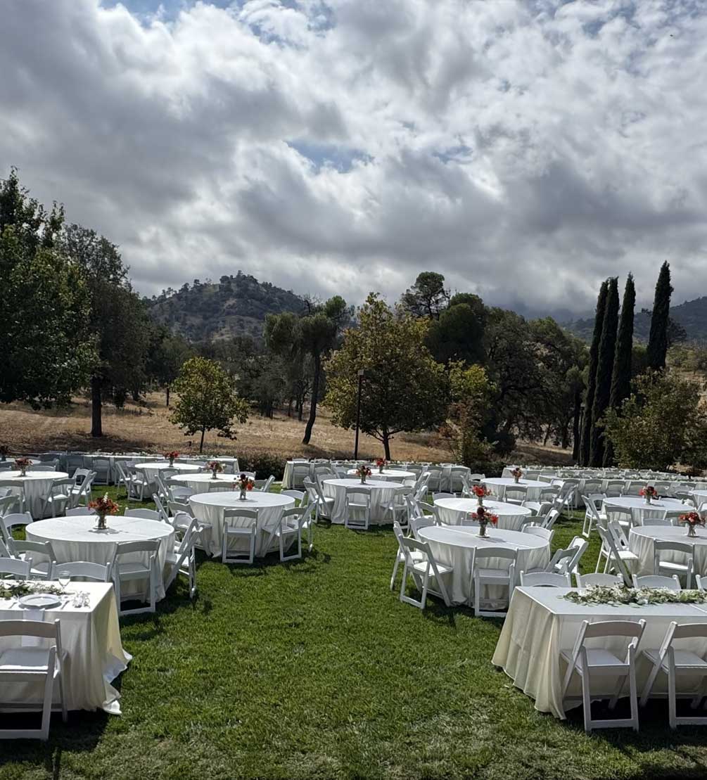 White-draped banquet tables set on the lawn beneath dramatic clouds and Tehachapi foothills