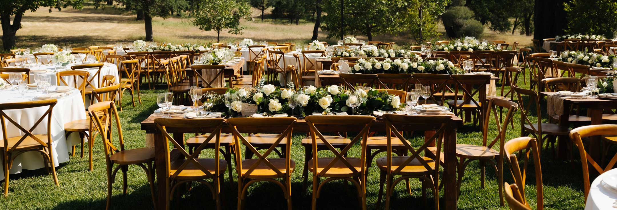 Wooden cross-back chairs and farm tables with white floral garlands at an outdoor reception