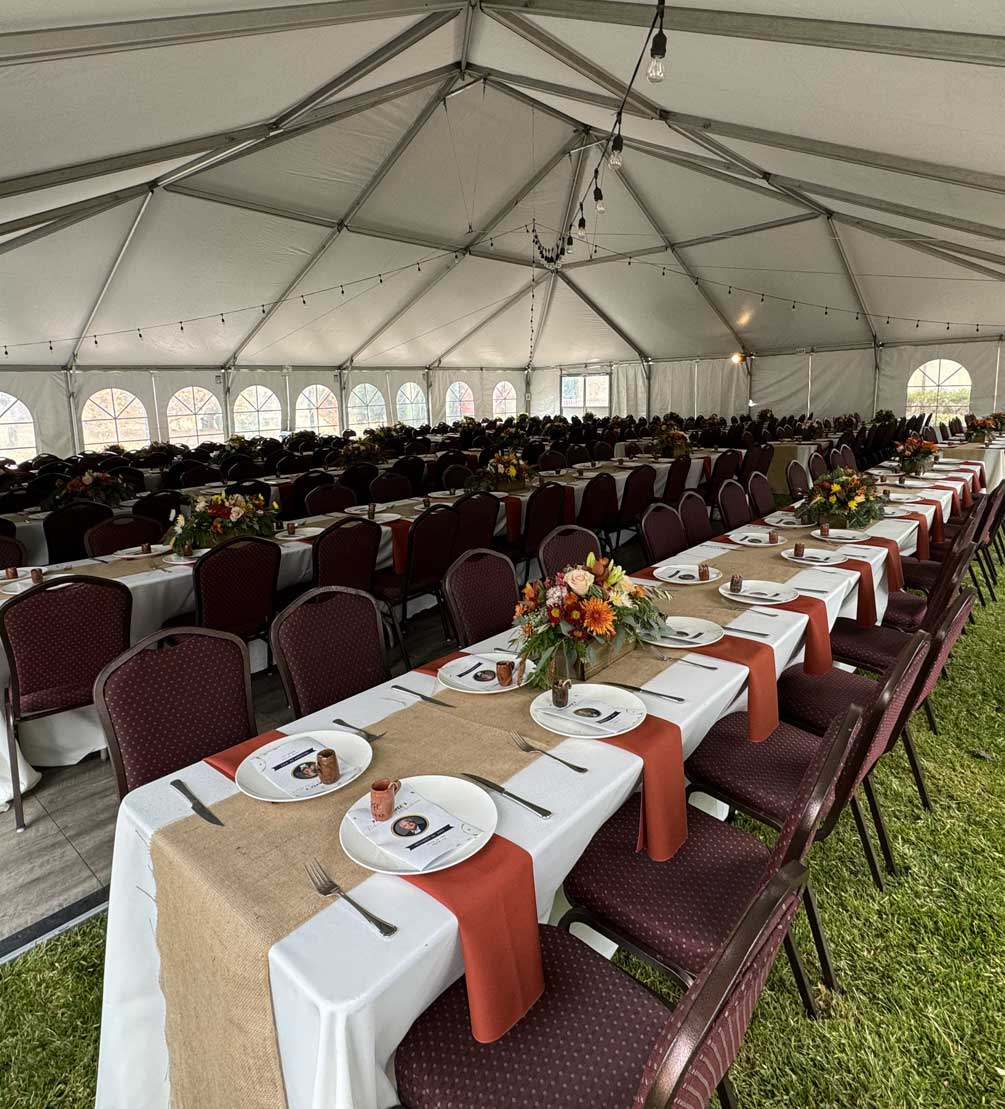 Rows of banquet tables with burlap runners, rust napkins, and floral centerpieces inside a large tent