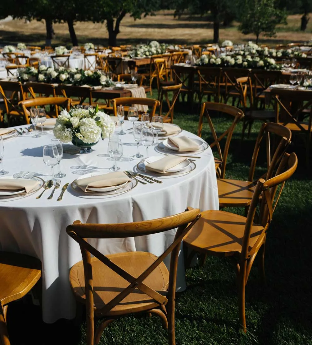 Round reception table with white linens, hydrangea centerpiece, and wooden cross-back chairs on the lawn