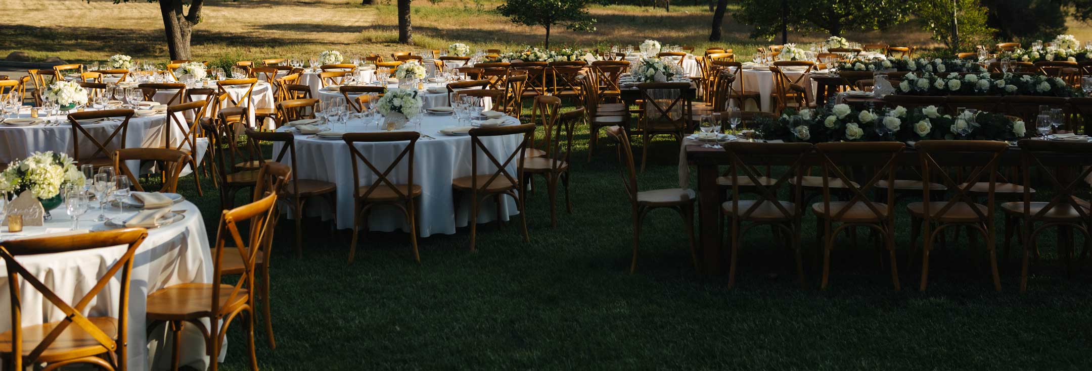 Outdoor wedding reception with round white-linen tables and wooden cross-back chairs under oak trees