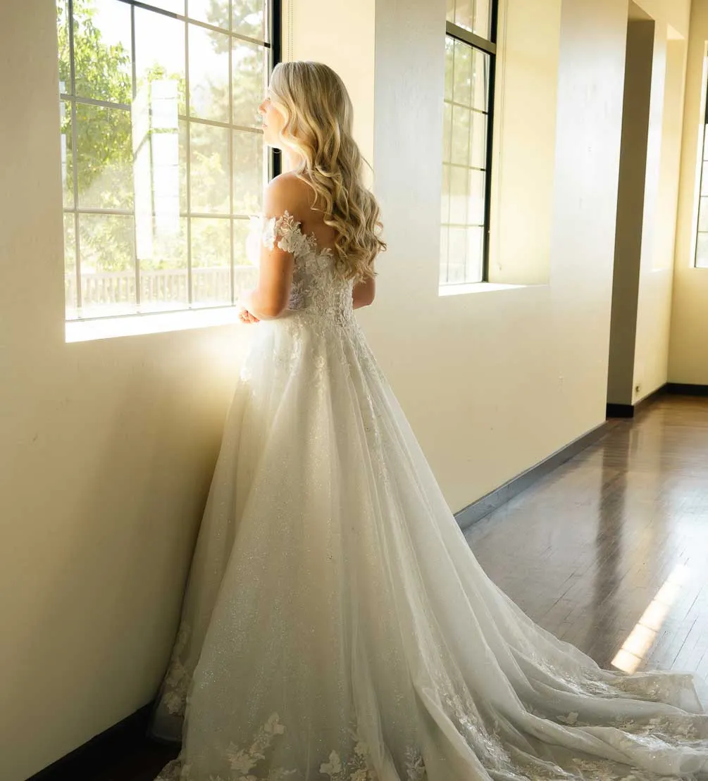 Bride in a lace gown gazing out a tall sunlit window in a wood-floored hall