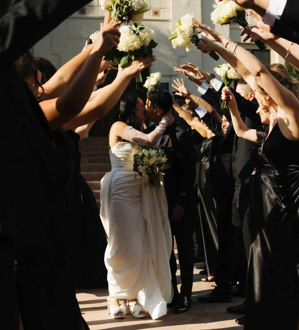 Bride and groom kissing as their wedding party raises white bouquets overhead