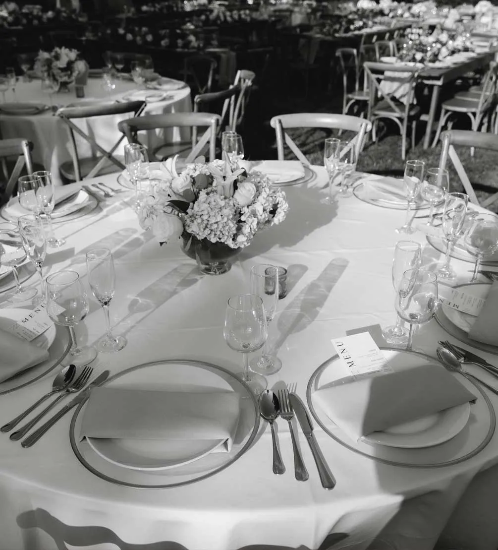 Black-and-white photo of a round reception table set with floral centerpiece, folded napkins, and menu cards