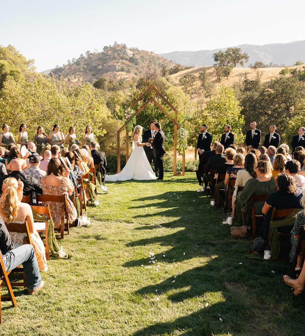 Couple exchanging vows under a wooden arch at an outdoor wedding ceremony framed by golden Tehachapi hills