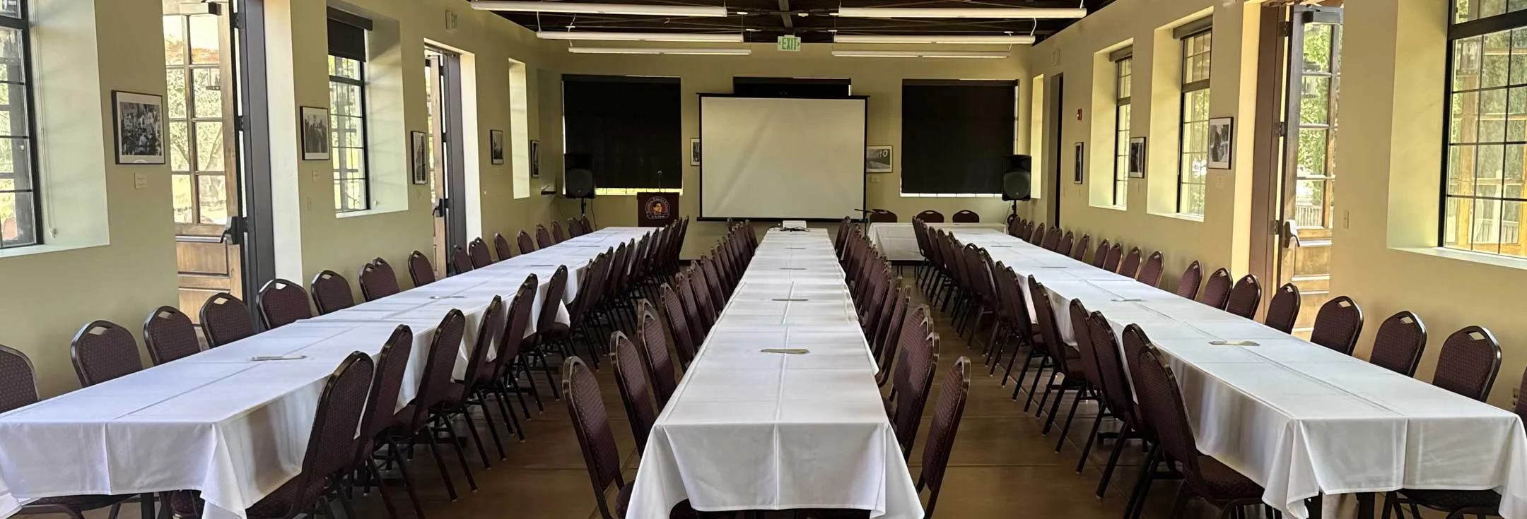 Conference hall set with long white-clothed banquet tables and chairs facing a projection screen