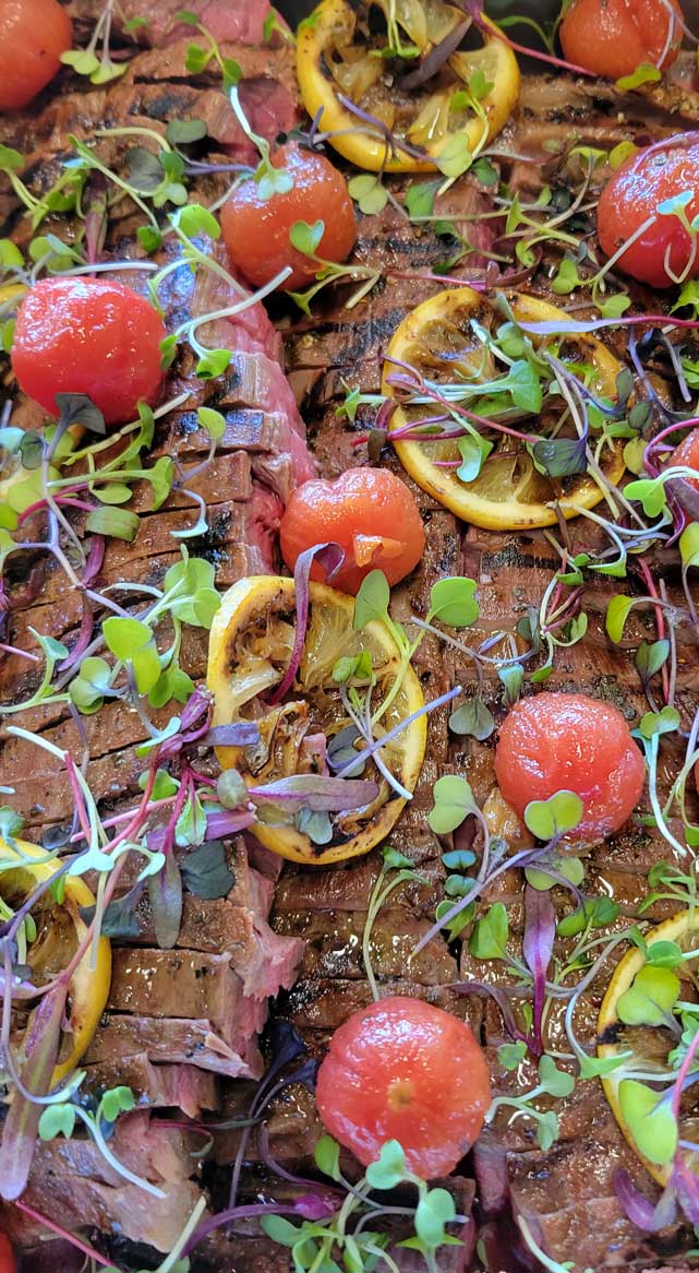 Sliced grilled steak garnished with charred lemon, blistered tomatoes, and microgreens