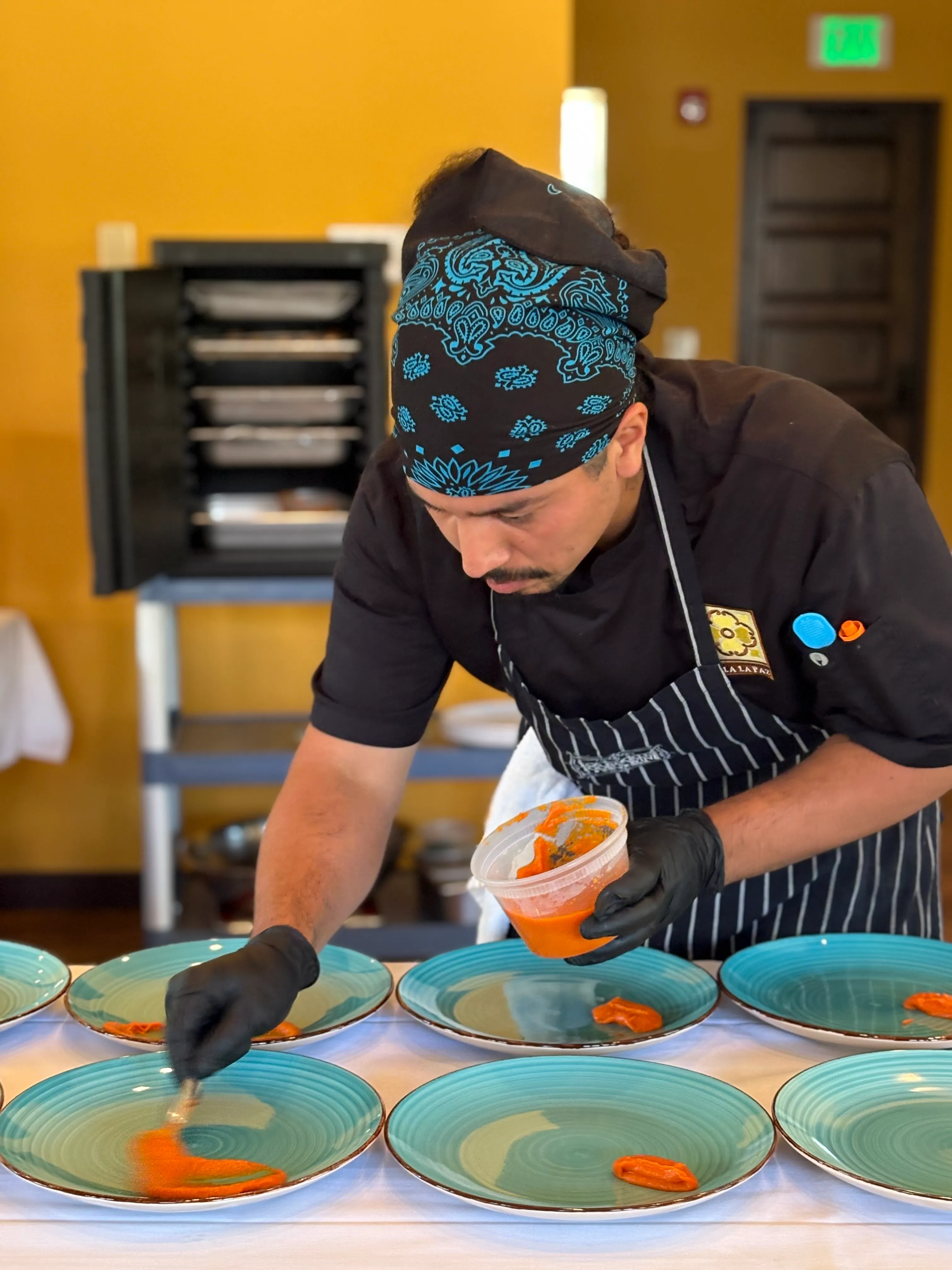 Chef in a bandana hand-saucing rows of turquoise plates while plating an event dinner