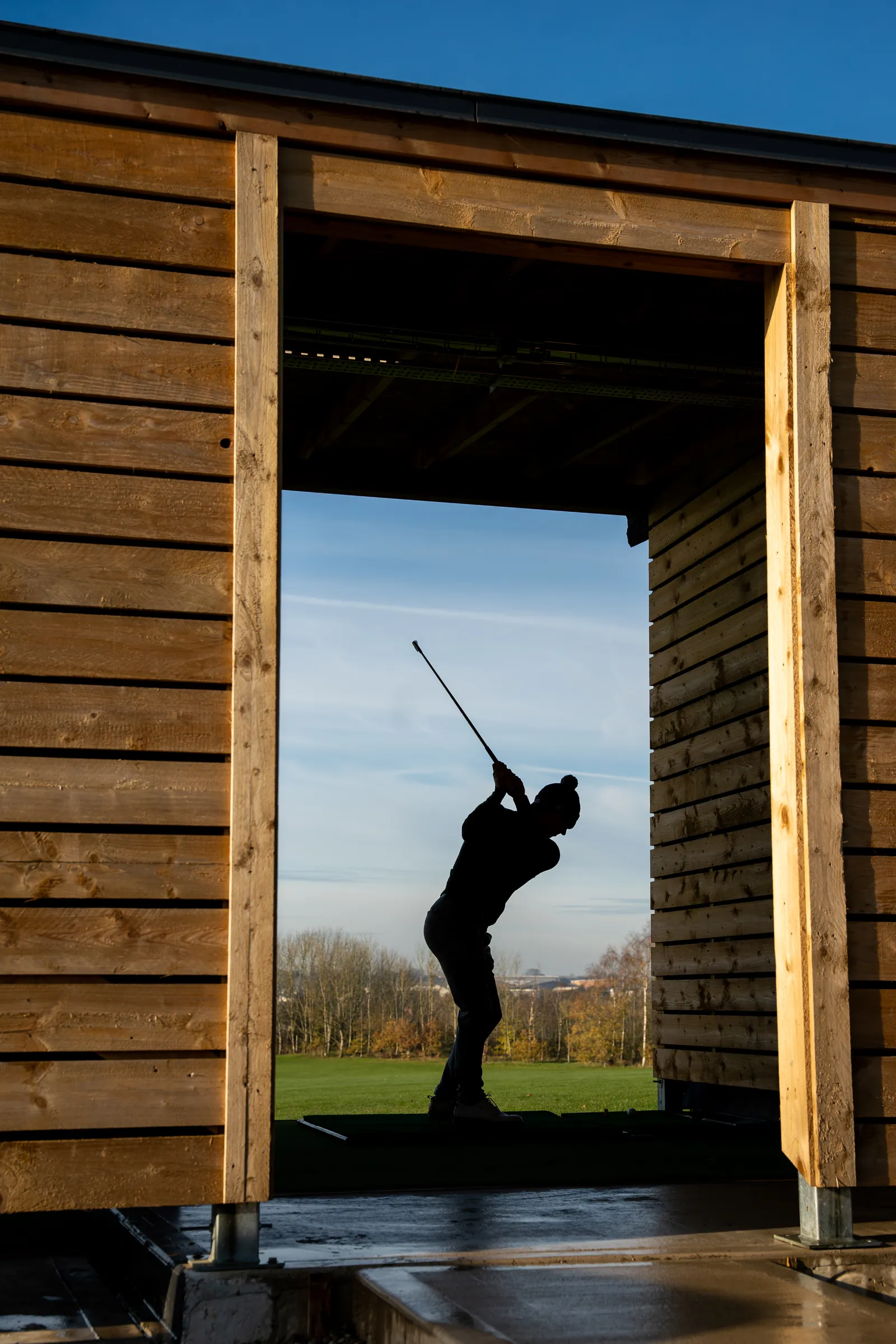 Silhouette of a golfer swinging at the open end of a driving range bay.