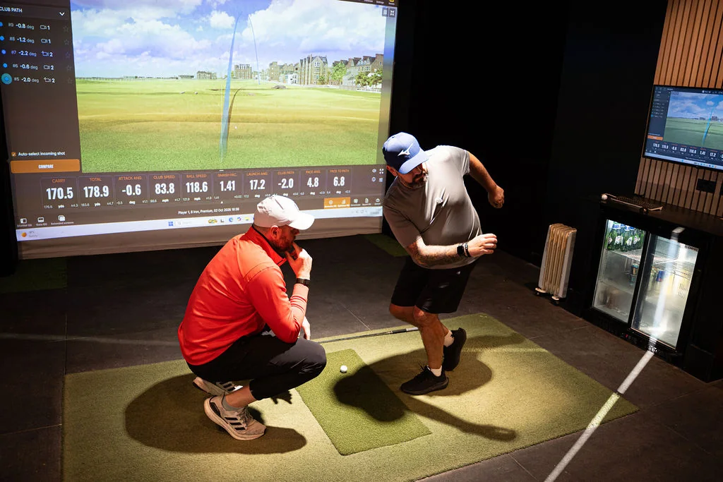 Coach crouching beside the hitting mat while a golfer steps through a movement assessment at Evolution Golf Academy near Derby